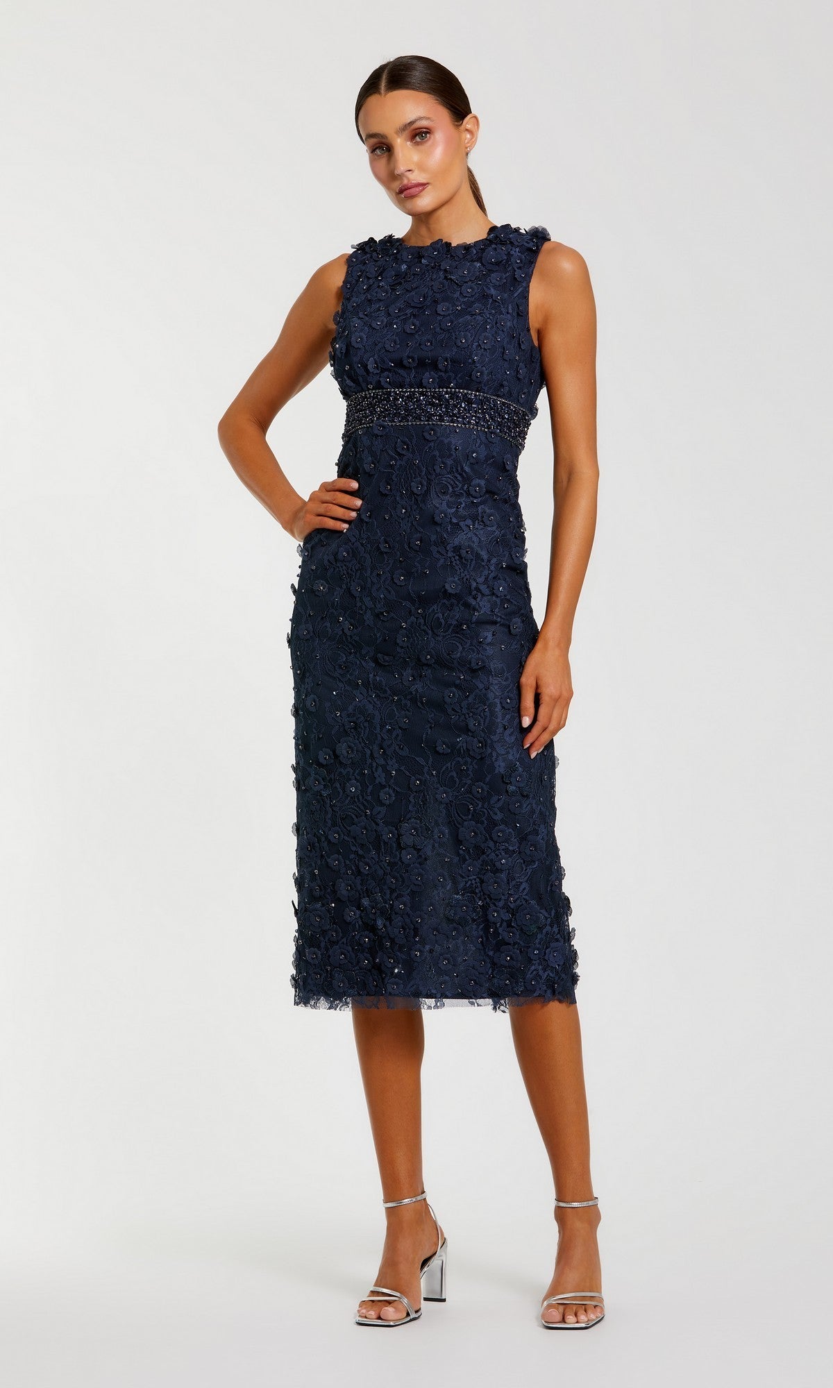A woman models the Mac Duggal 67853, a sleeveless navy blue knee-length party dress with floral details, paired with silver high-heeled sandals against a plain background.